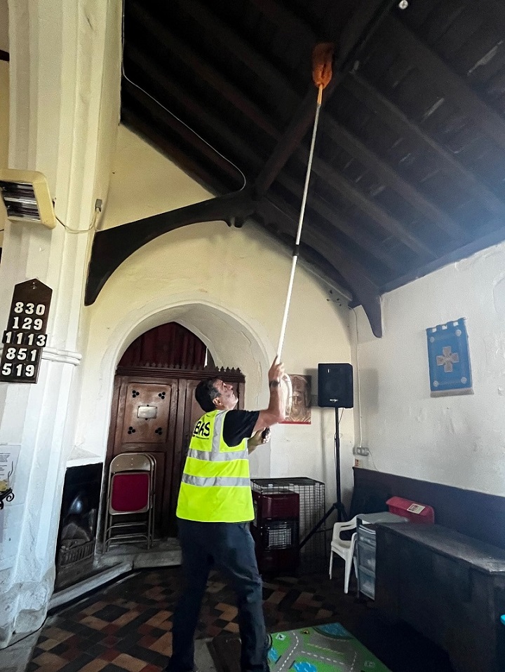 Image of volunteer in high vis clothing, with long duster cleaning the rafters in the church.