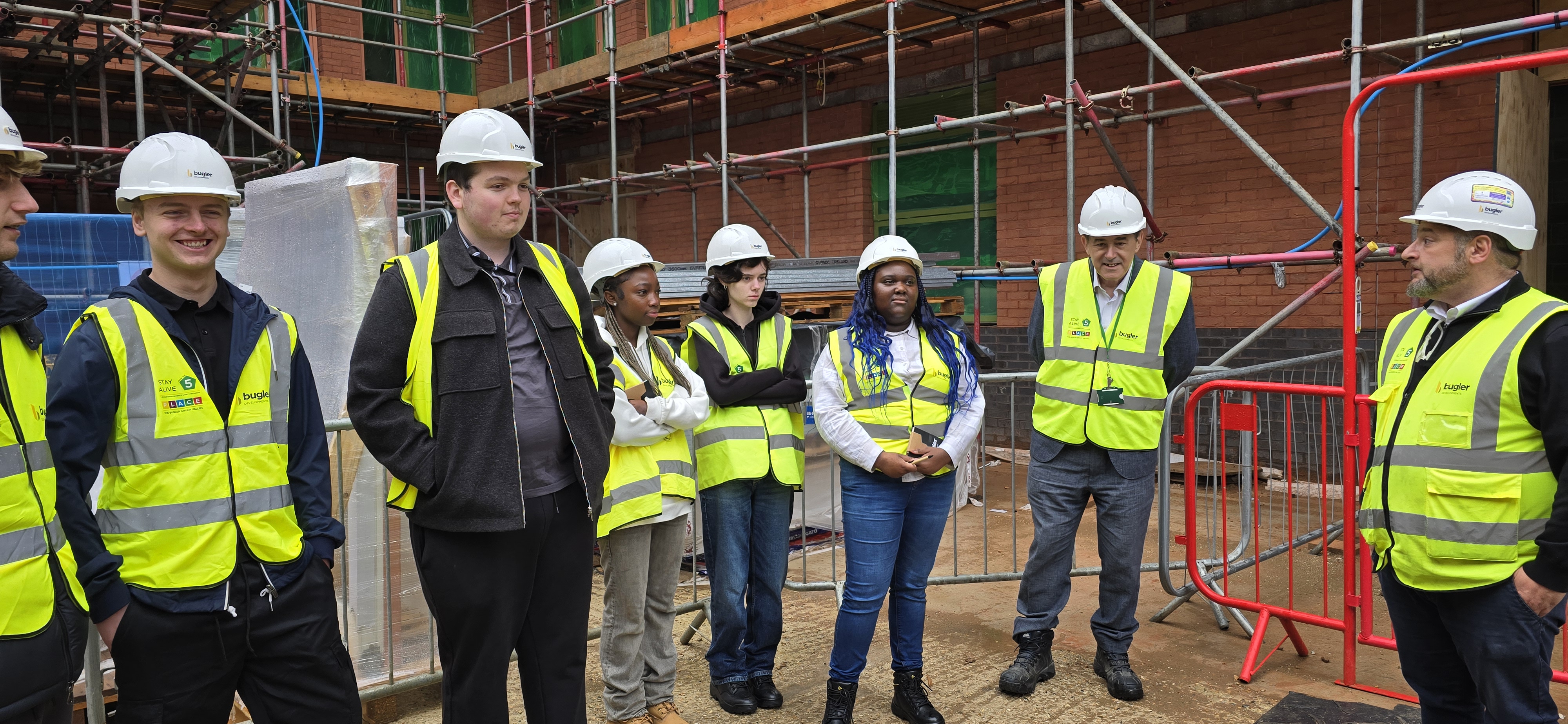 Image shows a group of people in high vis vests, hard hats, smiling and focusing on person speaking, on a building site