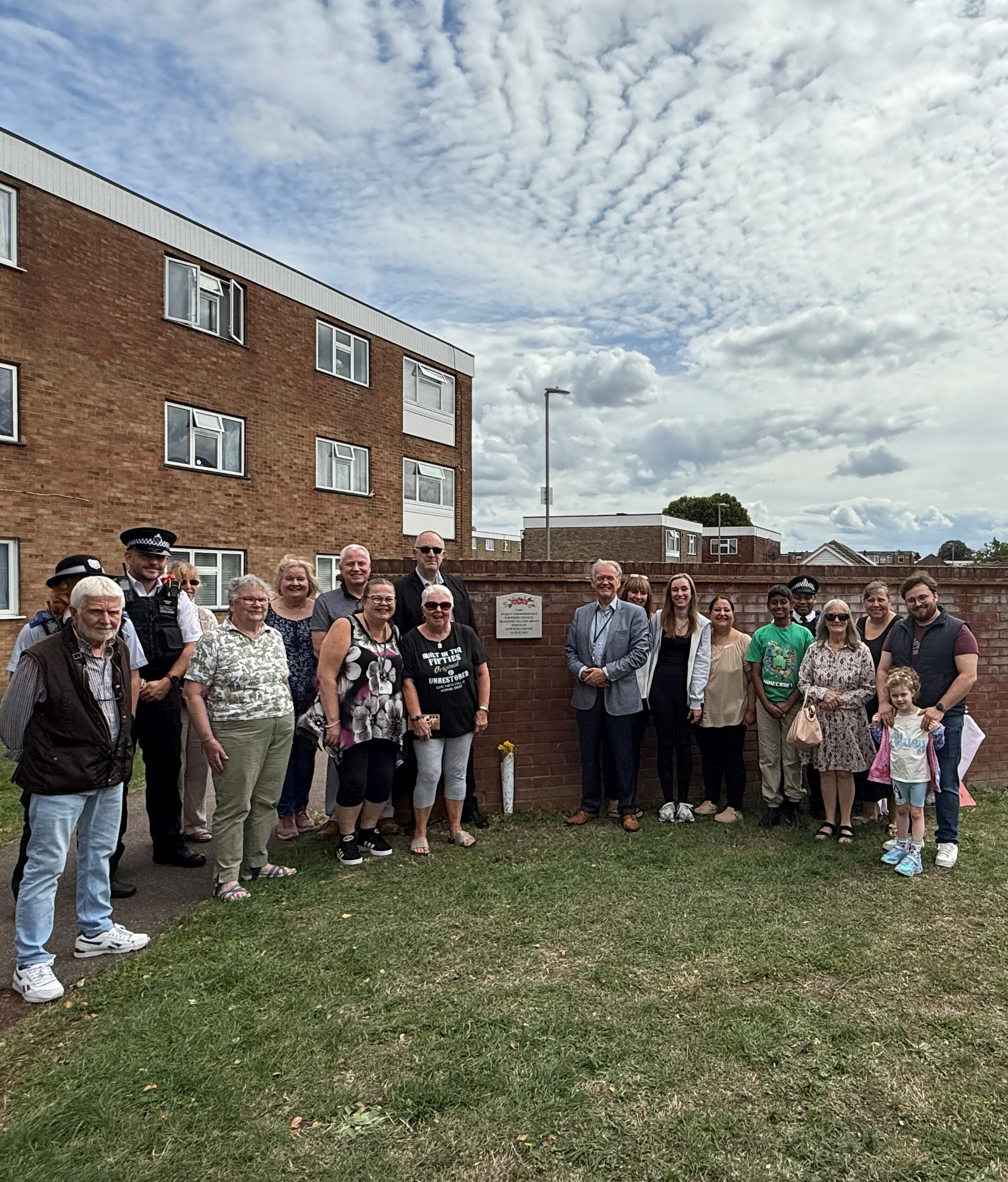 Image shows group smiling at camera, surrounding a wall plaque on a red brick wall