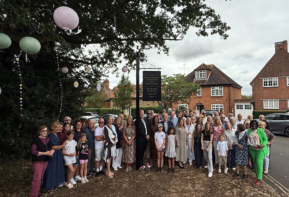 Image shows large group of people smiling at the camera next to a tall pole with a sign displaying the village green dedication information in gold cursive writing