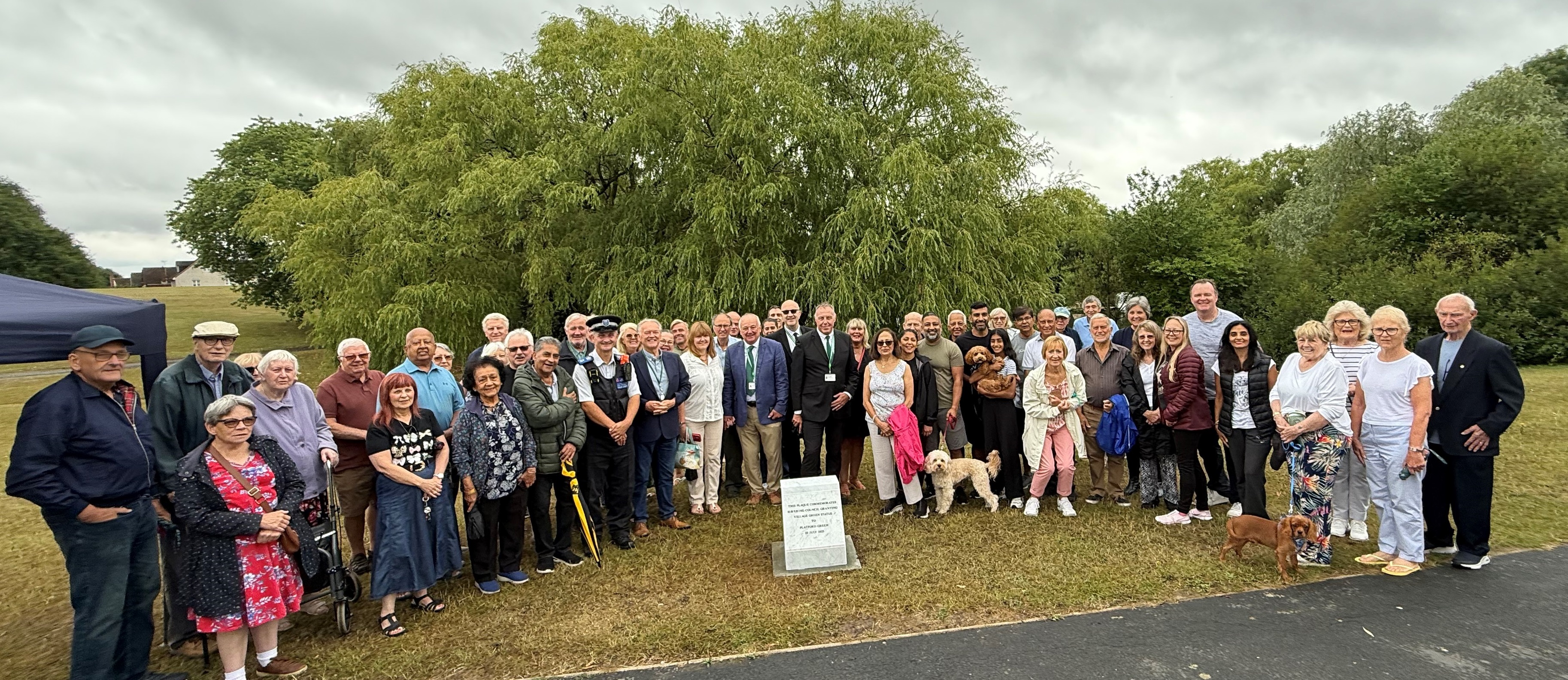 image shows large group of people in front of trees, smiling to the camera in front of a smalll white marble plinth